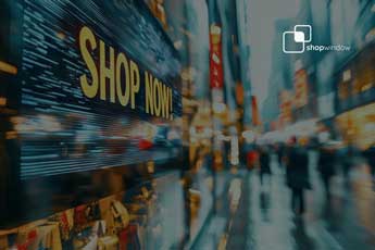 Schaufenster mit grosser «Shop Now!»-Aufschrift, daneben eine unscharfe, hell erleuchtete Einkaufsstrasse mit vorbeigehenden Passanten.